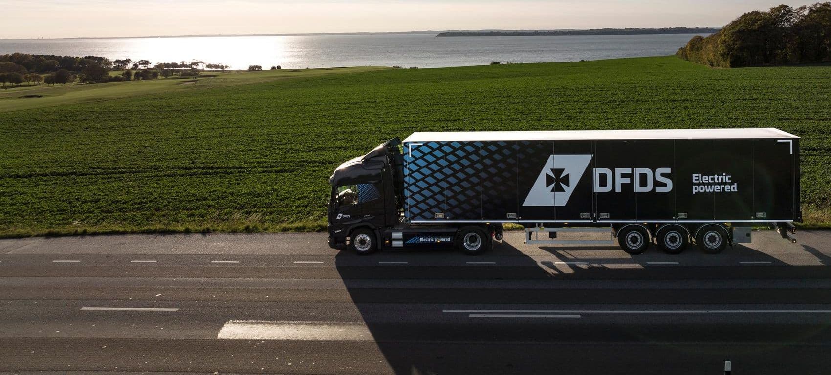 Blue electric truck with blue trailer driving on a road, with green surroundings