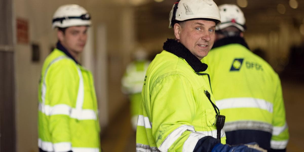 Three seamen, wearing white safety helmets and yellow safety clothing, walking inside a vessel, where two of them are turning around and into the camera