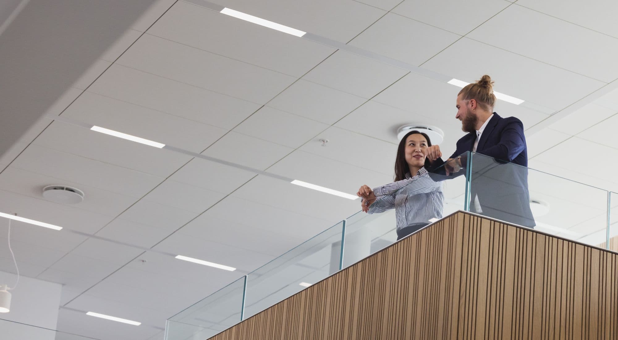 Two office workers, a man & woman, are talking to each other, leaning on the railing of a stairs platform in an office