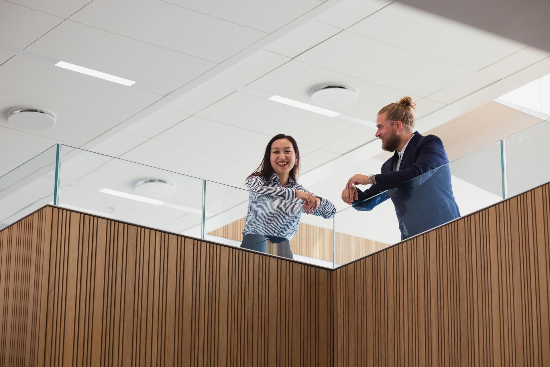 Two office workers, a man & woman, are talking to each other, leaning on the railing of a stairs platform in an office.