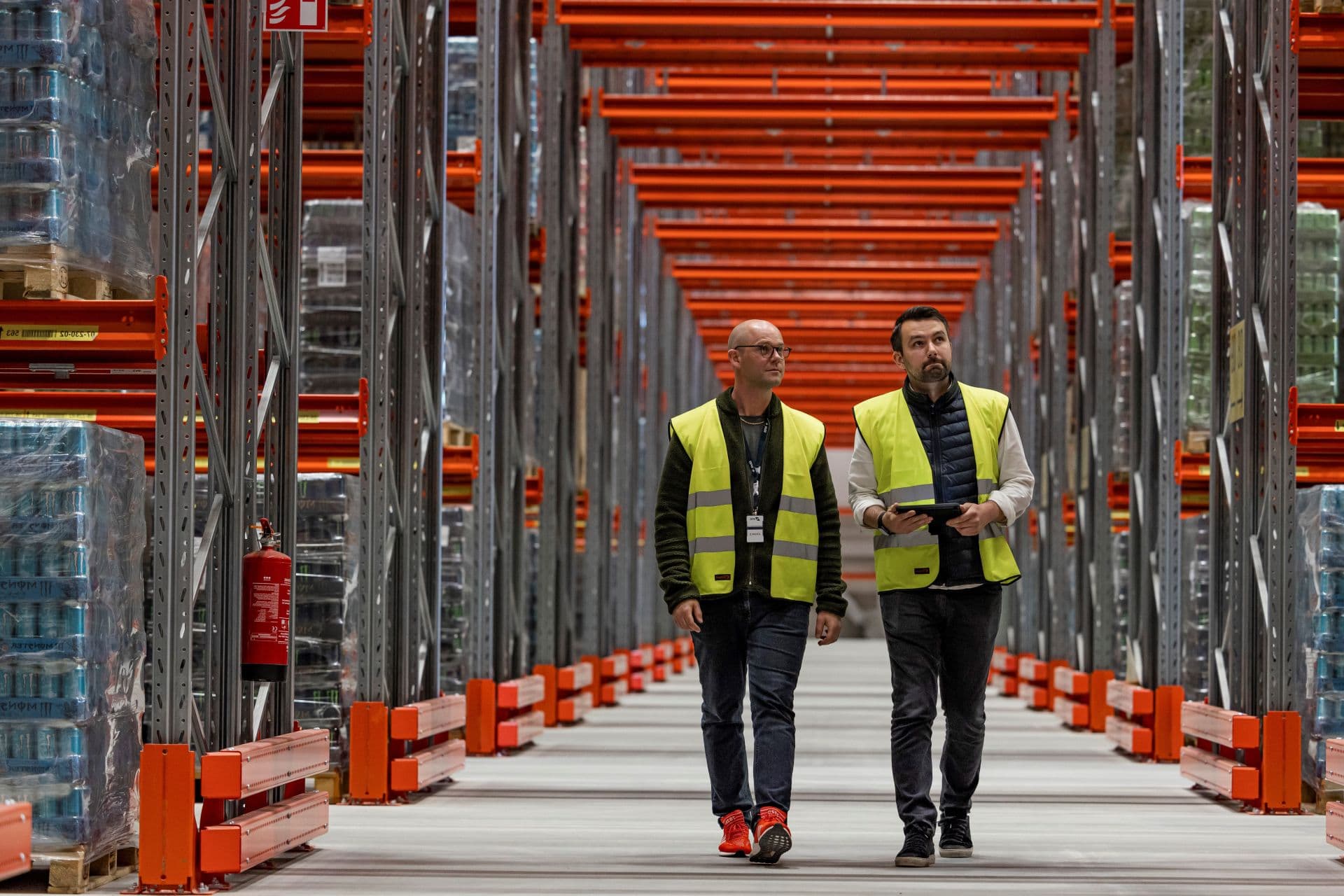 Two men wearing security vests walk in a hallway of a warehouse on both sides of gantries, loaded with pallets of goods