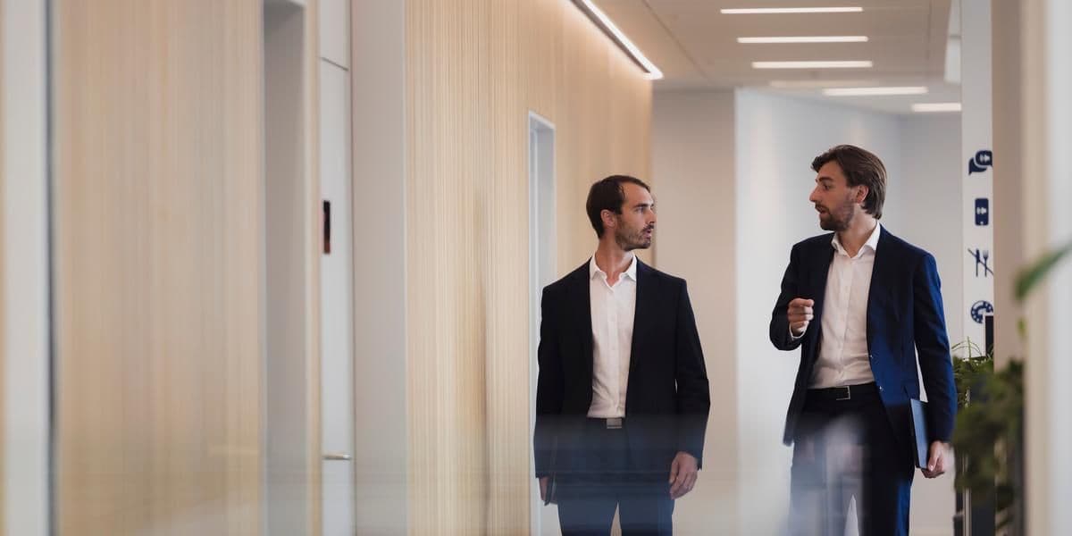 Two employees in dark suits, walking in a modern office, talking to each other