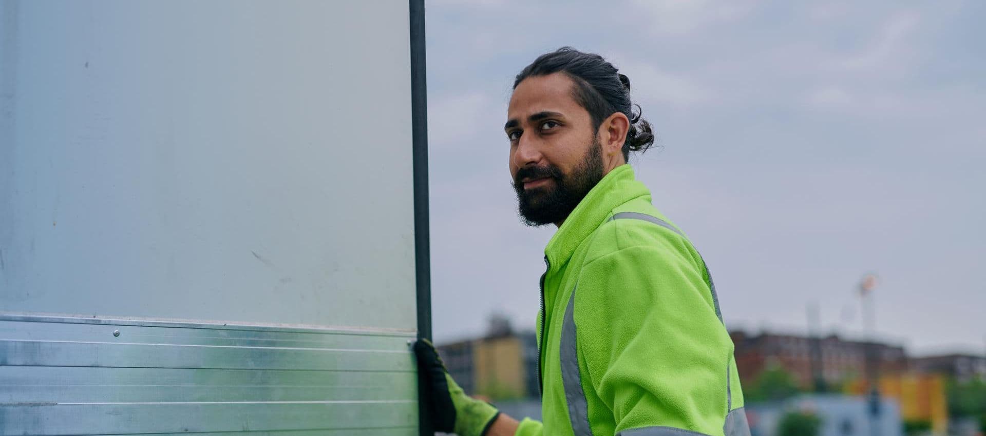 A male driver with a yellow security jacket, handling a door of a cold chain trailer, looking into the distance